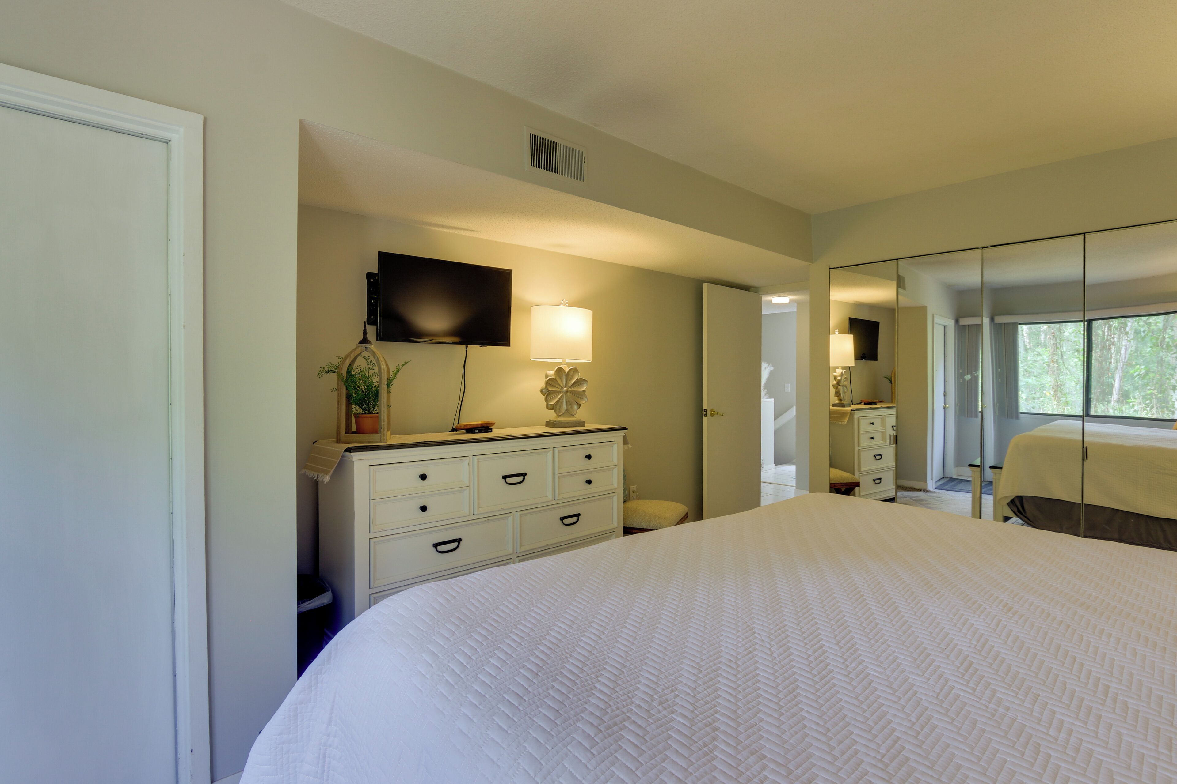 A hotel room with a large bed, a dresser with drawers, a flat-screen TV mounted on the wall, and a large mirror reflecting the room.