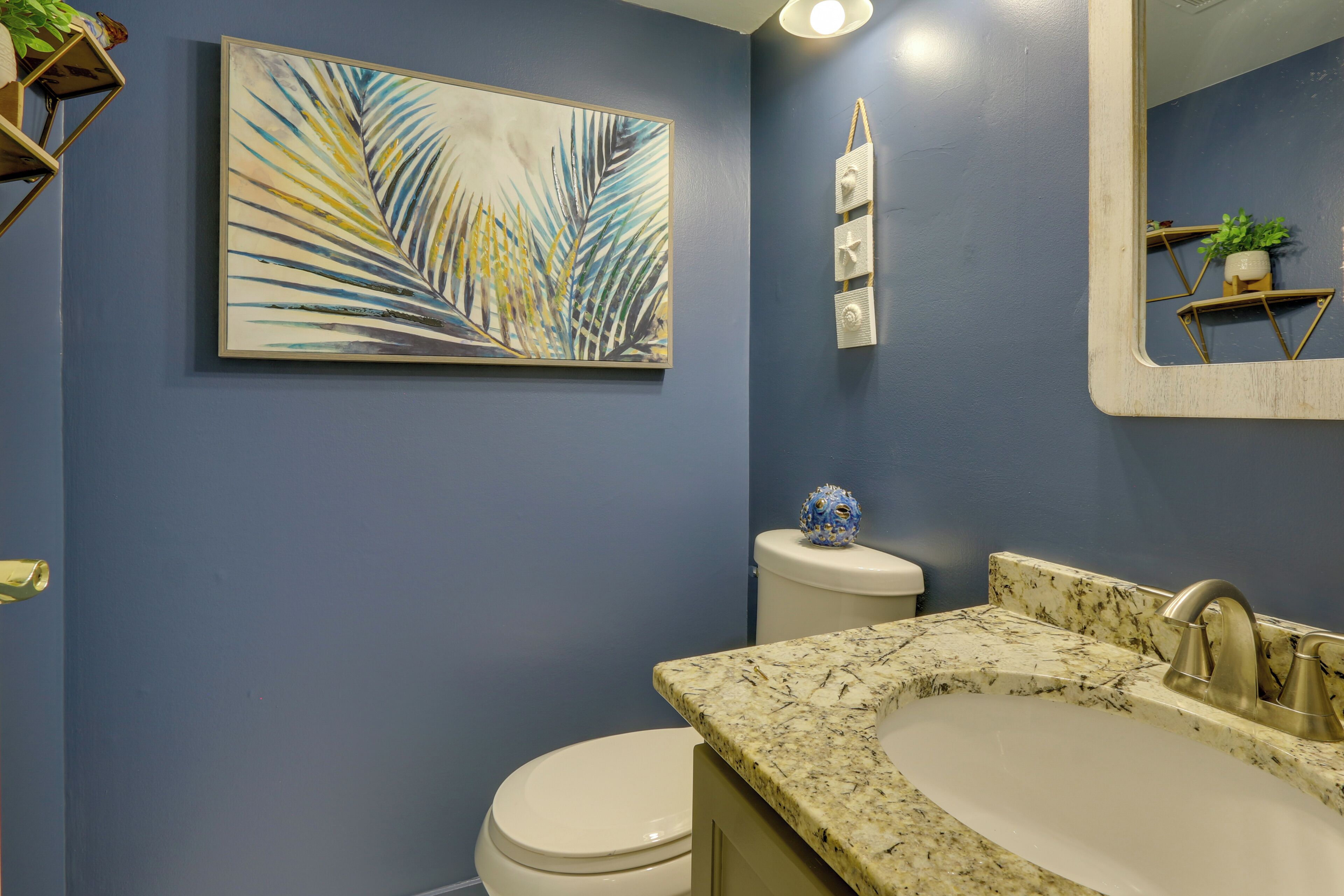 A bathroom with a blue wall, a white toilet, a marble sink, and a painting of palm leaves.