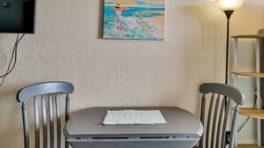 Une pièce avec un tableau représentant une femme au bord de la mer, une table avec un set de table, deux chaises, une lampe et une horloge au mur.
