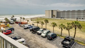 Un parking où sont garés différents véhicules, un immeuble à plusieurs étages et, au loin, une plage.