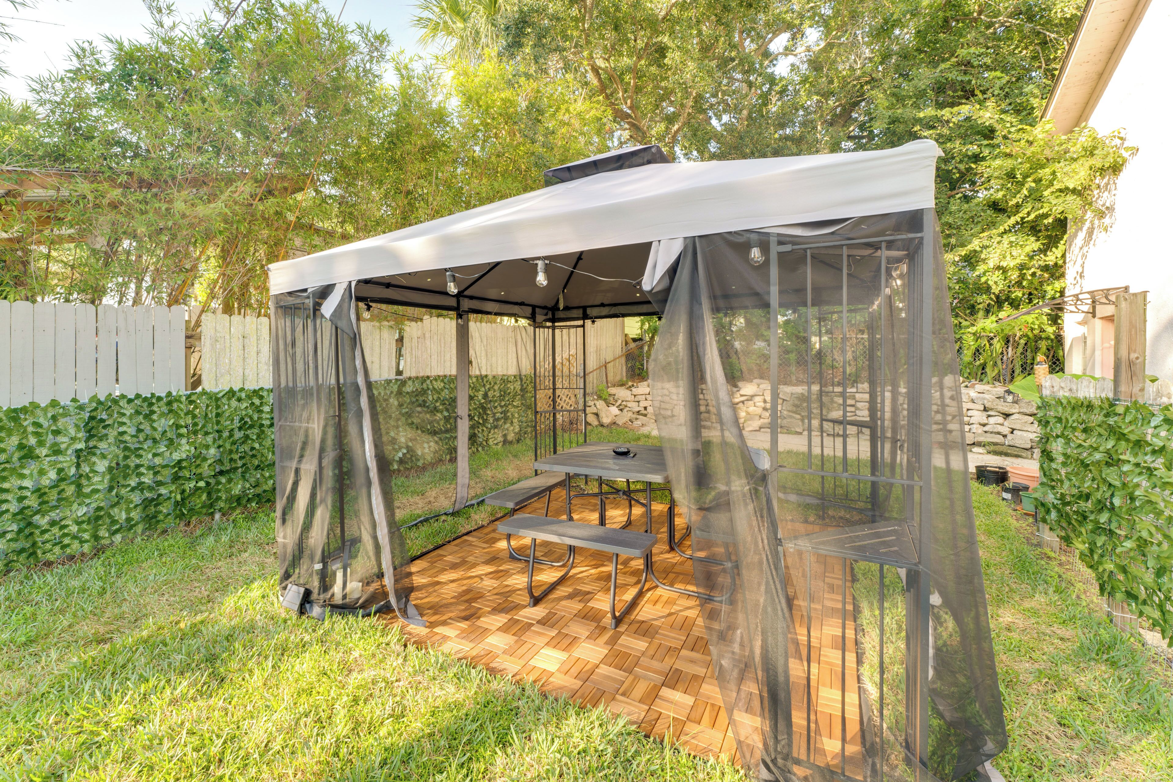 A covered outdoor seating area with a table and benches, surrounded by a wooden deck and a white fence.