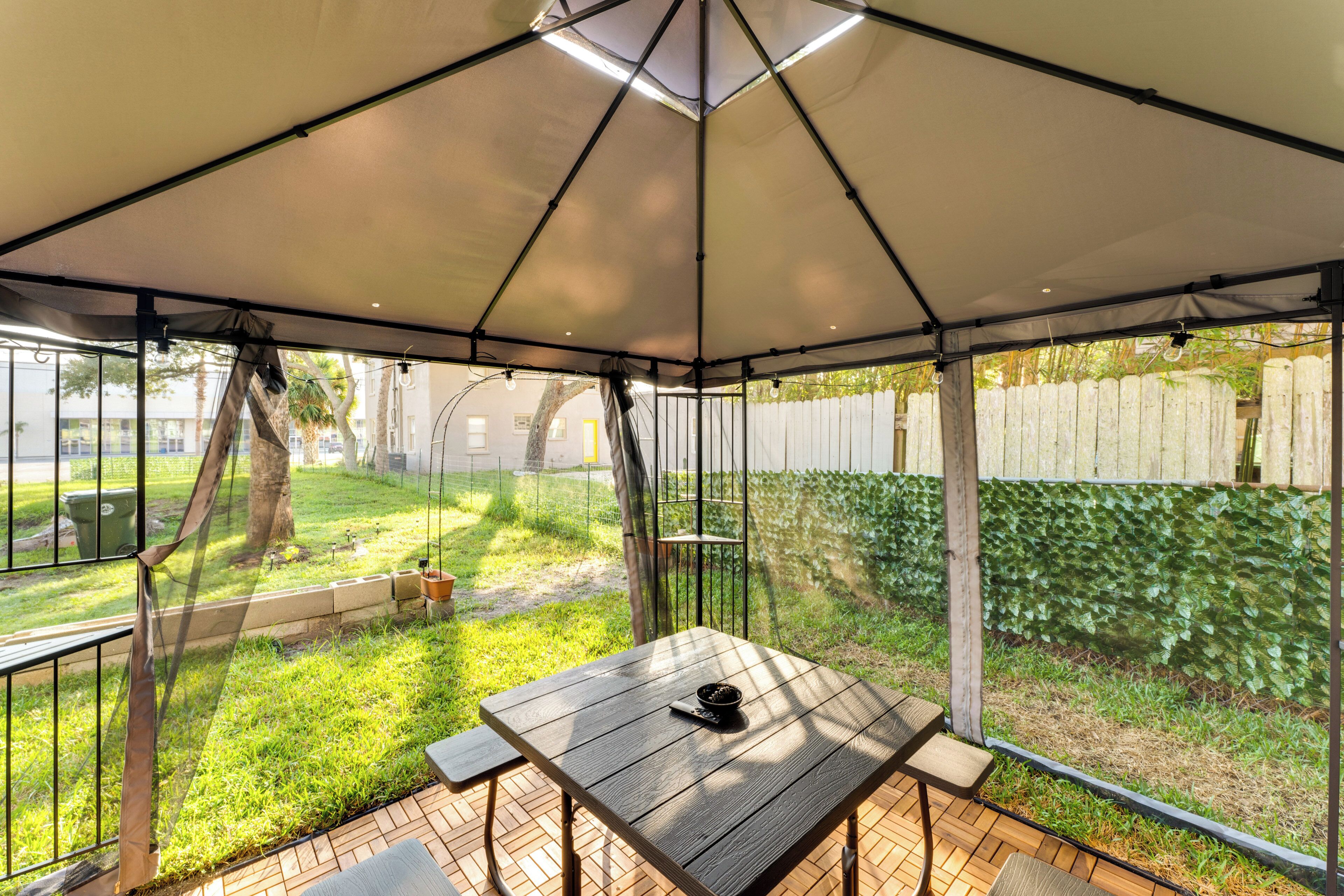 Un patio techado con mesa y sillas, rodeado de un jardín y una cerca.