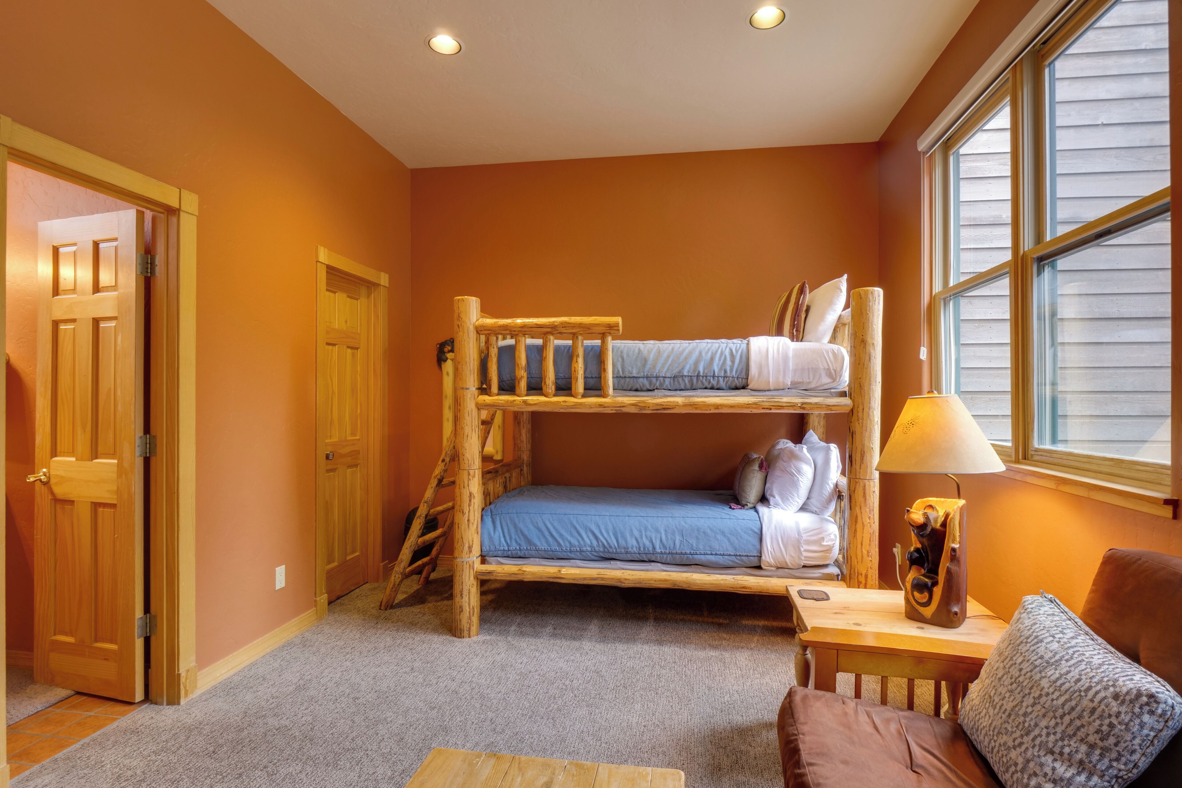 A bunk bed room with a wooden ladder, a small table with a lamp, and a window with blinds.