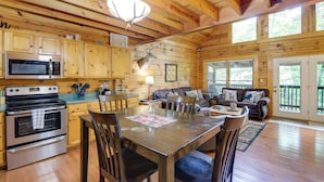 A cabin interior with wooden walls, a kitchen with a microwave and oven, a dining table with chairs, and a living area with a couch and fireplace. - Hot Tub + Pool Table: Secluded Seiverville Cabin (Sevierville)