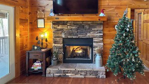 A cozy living room with a stone fireplace, a Christmas tree, and a flat-screen TV mounted on the wall. - Hot Tub + Pool Table: Secluded Seiverville Cabin (Sevierville)