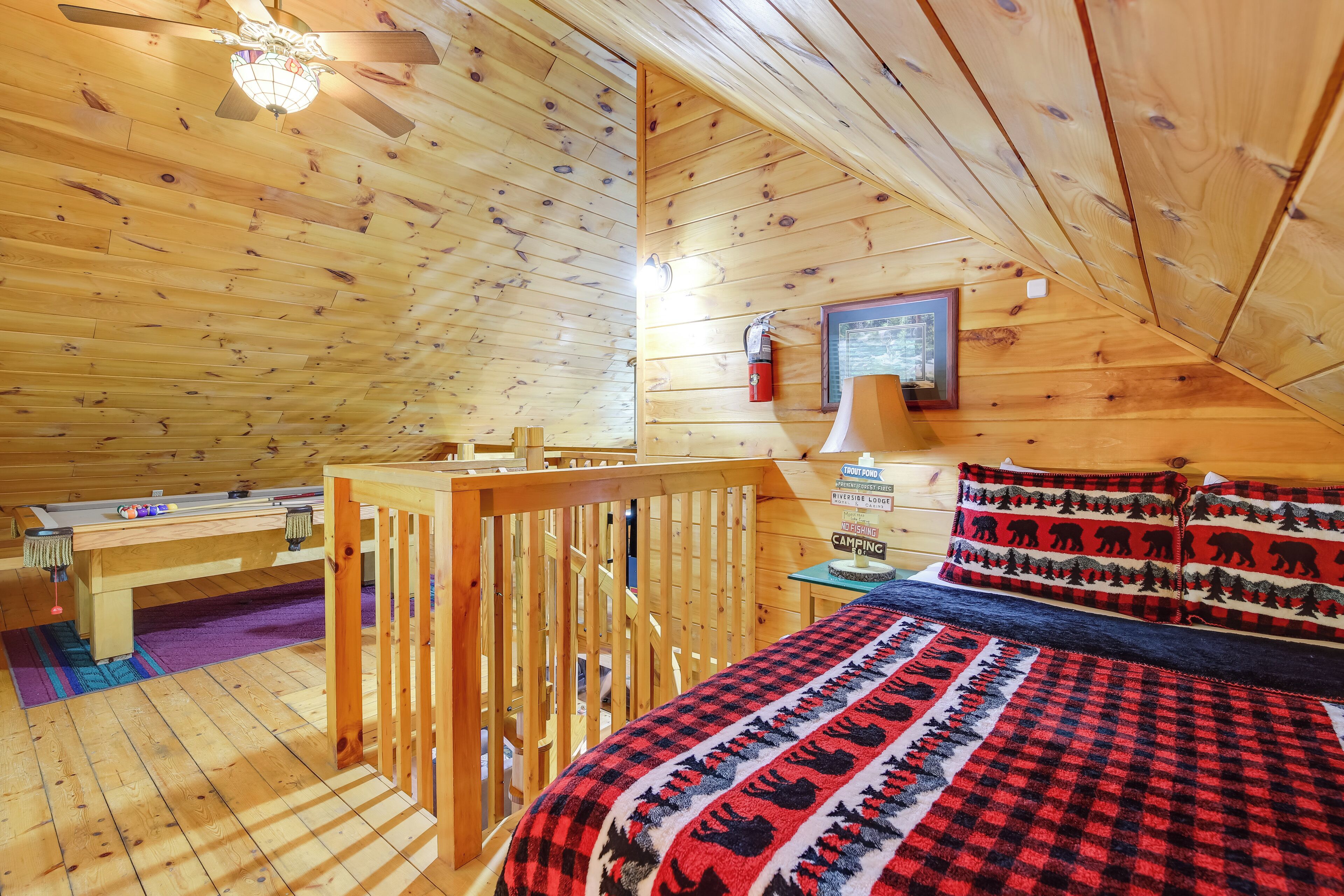 A cozy cabin interior with a wooden ceiling, a bed with a red and black checkered blanket, a small table, and a ceiling fan.