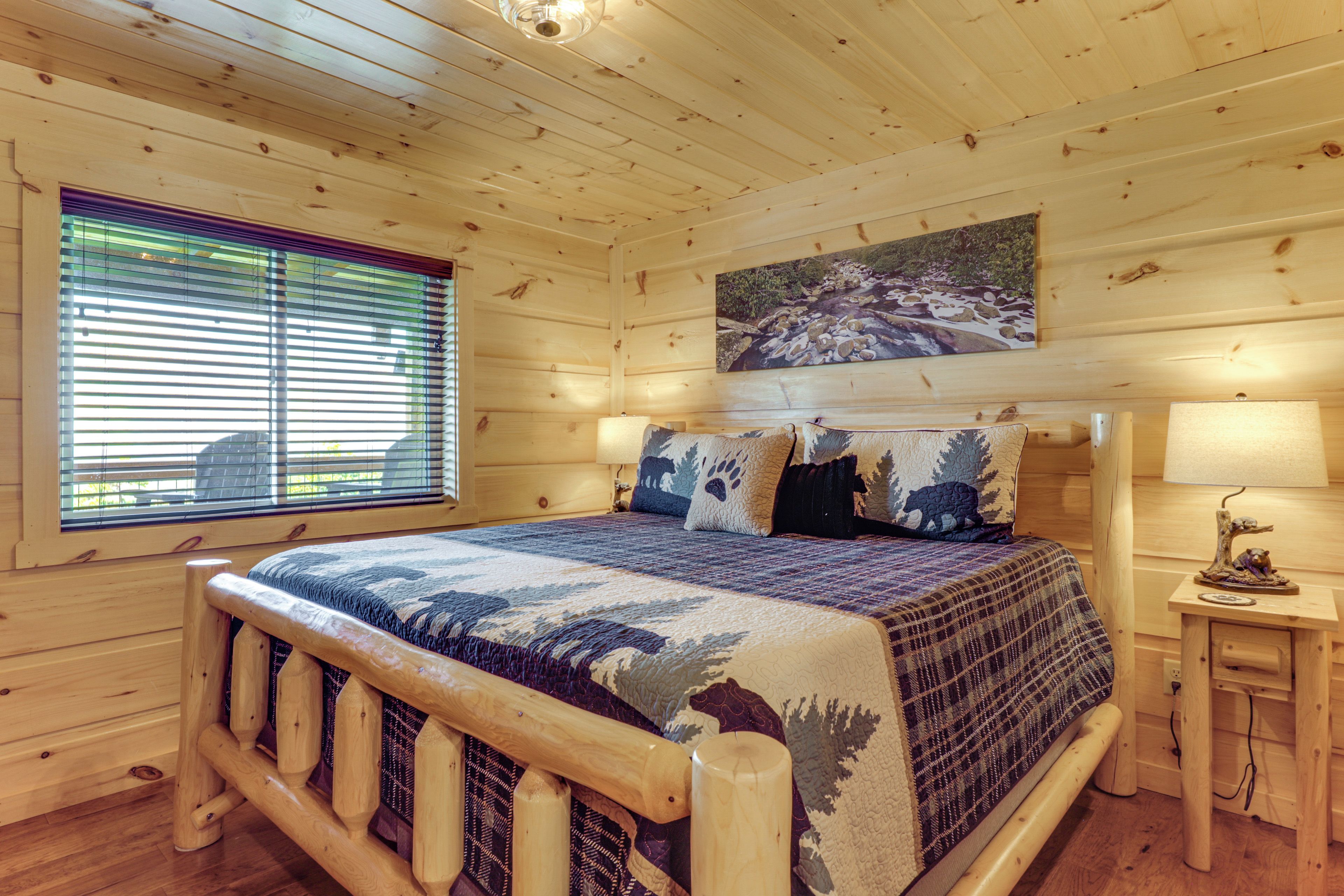 Un dormitorio de cabaña de madera con una cama, una ventana con persianas, un cuadro sobre la cama y una mesita de noche con una lámpara.