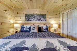 A wooden cabin bedroom with a bed, two lamps, a painting of a stream, and a door. - Gorgeous Gatlinburg Cabin: Large Deck & Hot Tub! (Gatlinburg)