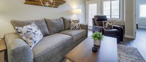 A living room with a gray sofa, a wooden coffee table, and a black leather chair. There is a wall-mounted star decoration and a 'thankful' sign above the door.