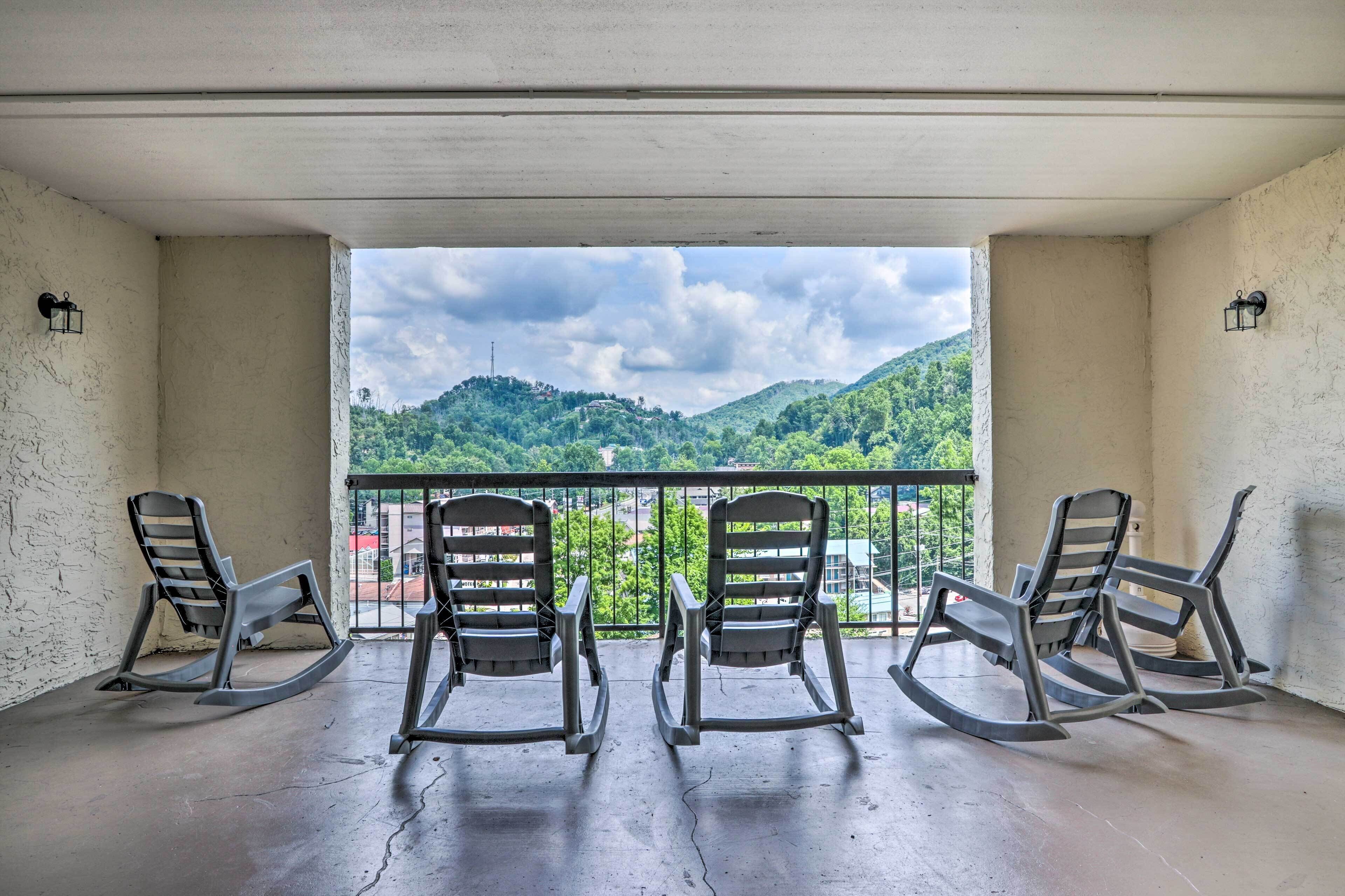A balcony with three rocking chairs facing a scenic view of green hills and a cloudy sky.