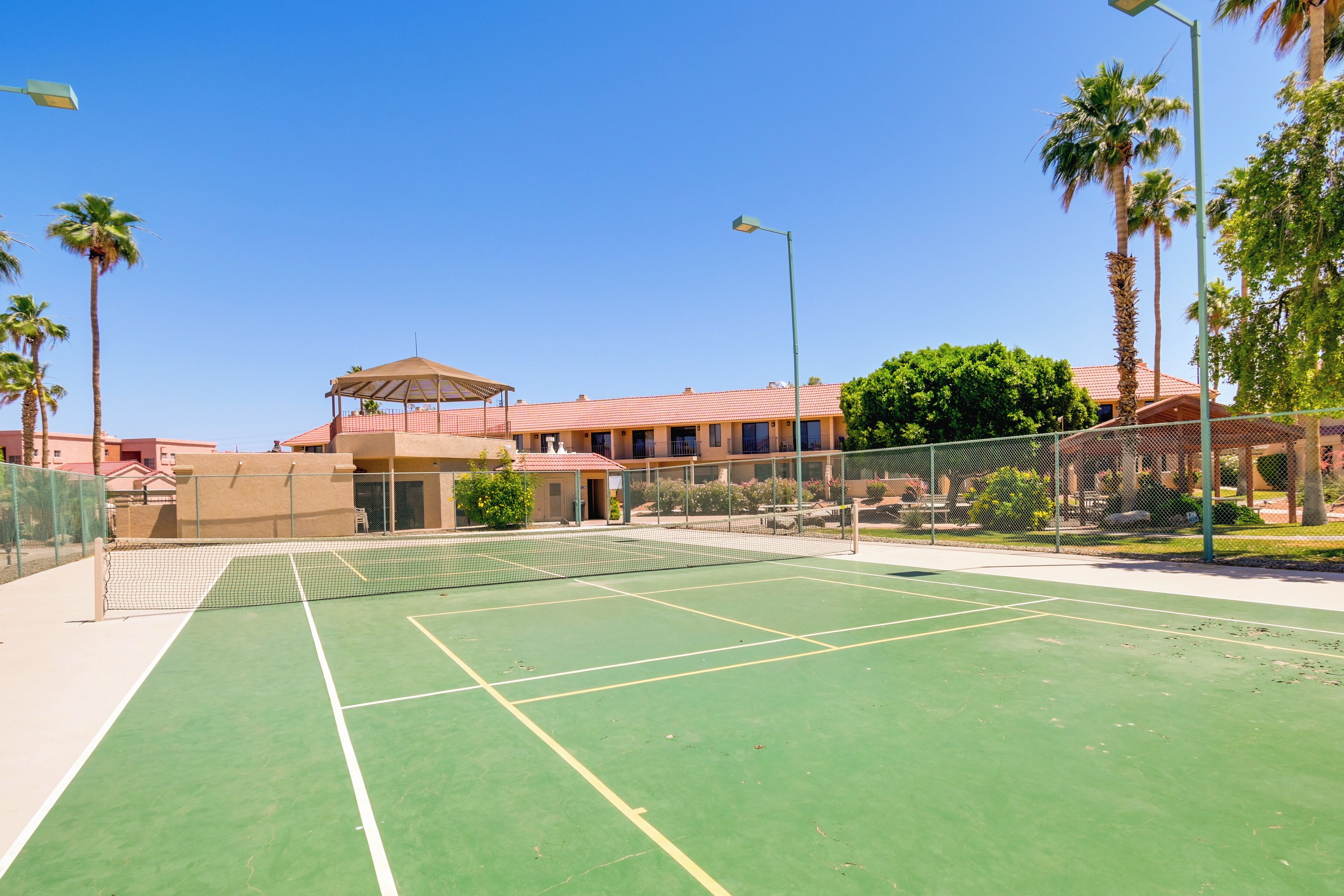 Una cancha de tenis con una red, rodeada por una cerca y palmeras.