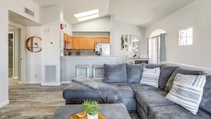 A modern living room with a gray sectional sofa, a wooden coffee table with a plant, and a kitchen in the background with wooden cabinets and a white refrigerator. - Walk to Rotary Park: Lake Havasu City Condo (Lake Havasu City)