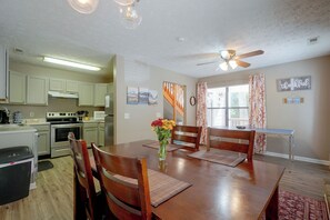 Un comedor con una mesa de madera, sillas y un jarrón de flores. Se observa una cocina al fondo con armarios y electrodomésticos.