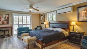 A spacious bedroom with a large bed, a desk, and a chair. There is a ceiling fan, an air conditioning unit, and a window with blinds.