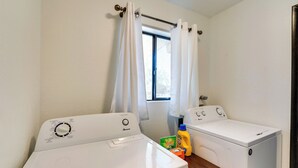 A laundry room with two white front-loading washers and dryers, a wooden table, and a window with white curtains.