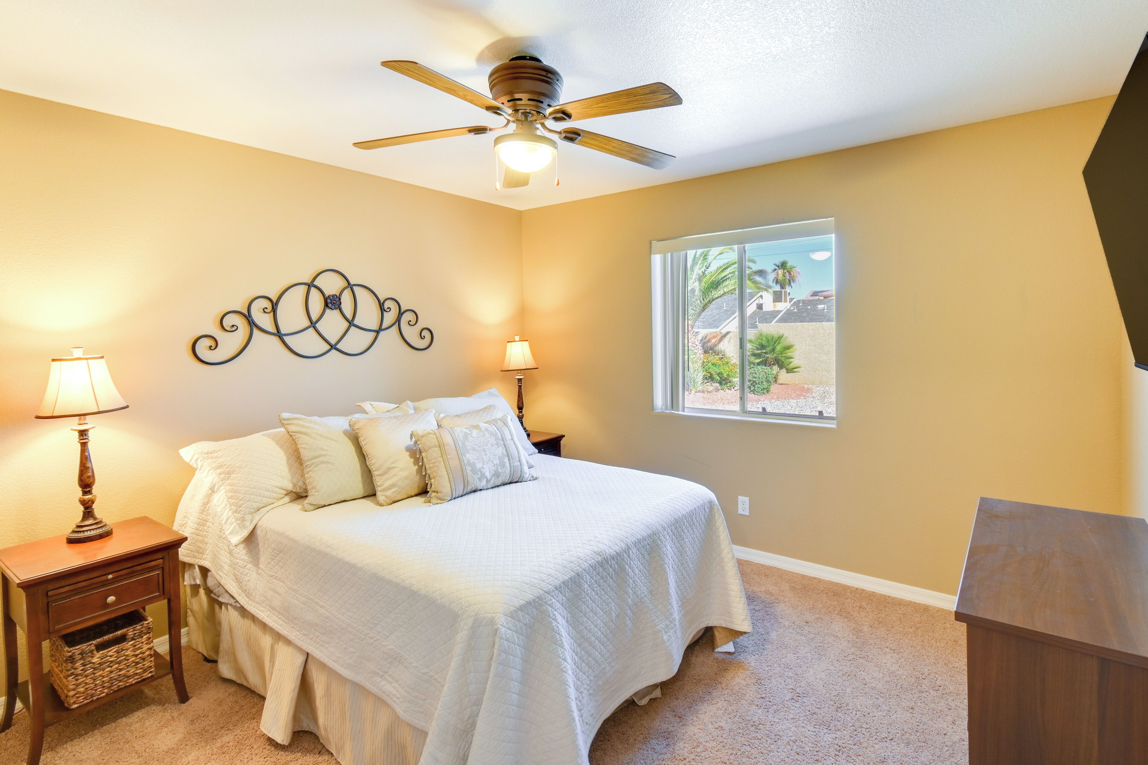 A bedroom with a bed, two bedside lamps, a ceiling fan, a window with a view, and a small wooden nightstand with a basket.