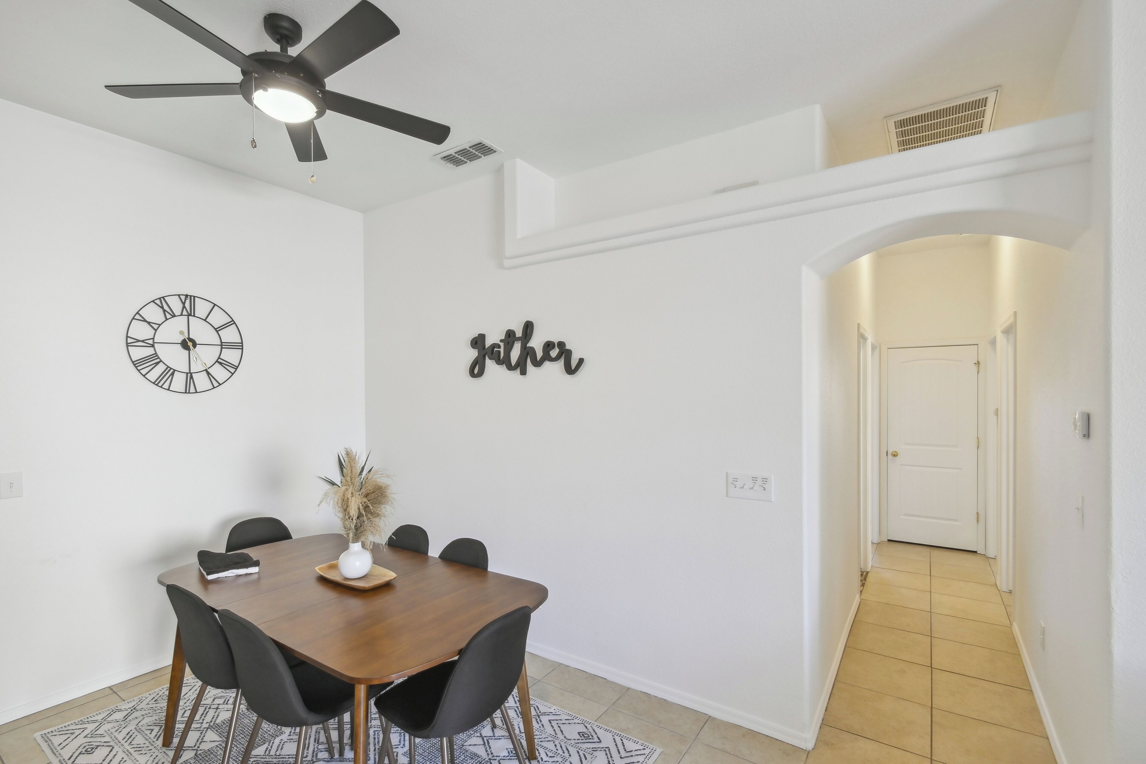 A dining area with a wooden table, black chairs, a ceiling fan, a wall clock, and a decorative wall piece.