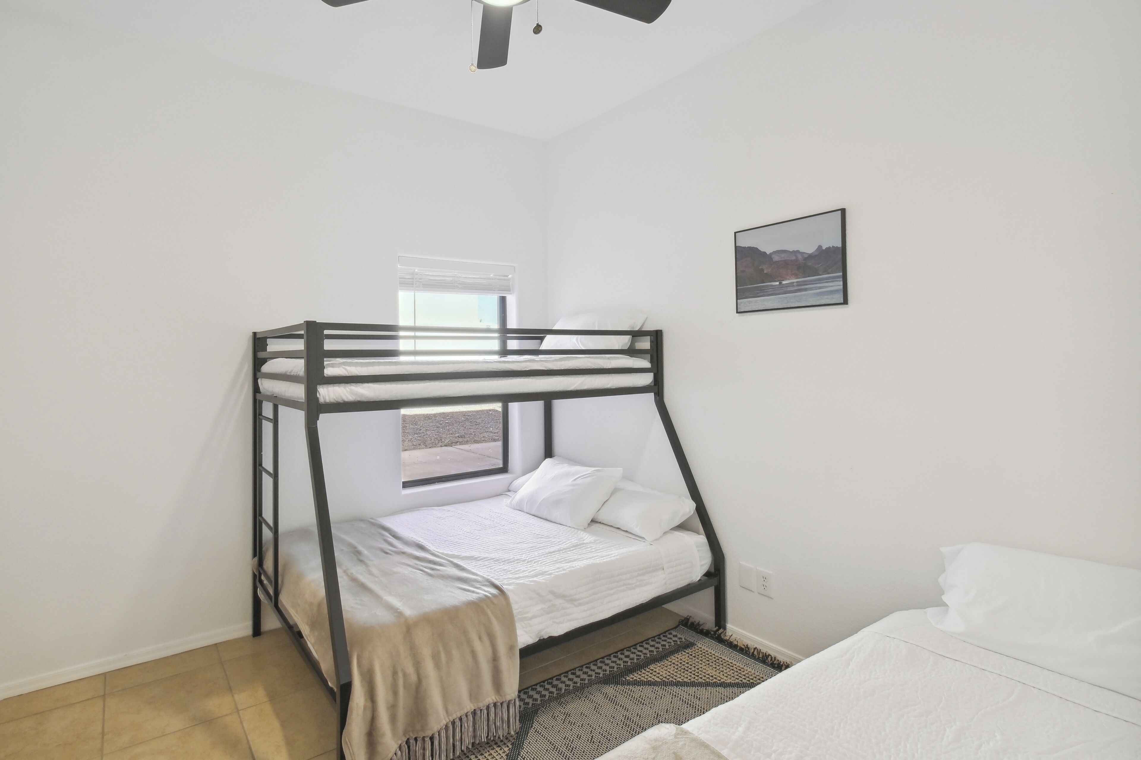 A bunk bed room with a window, a ceiling fan, and a framed picture on the wall.