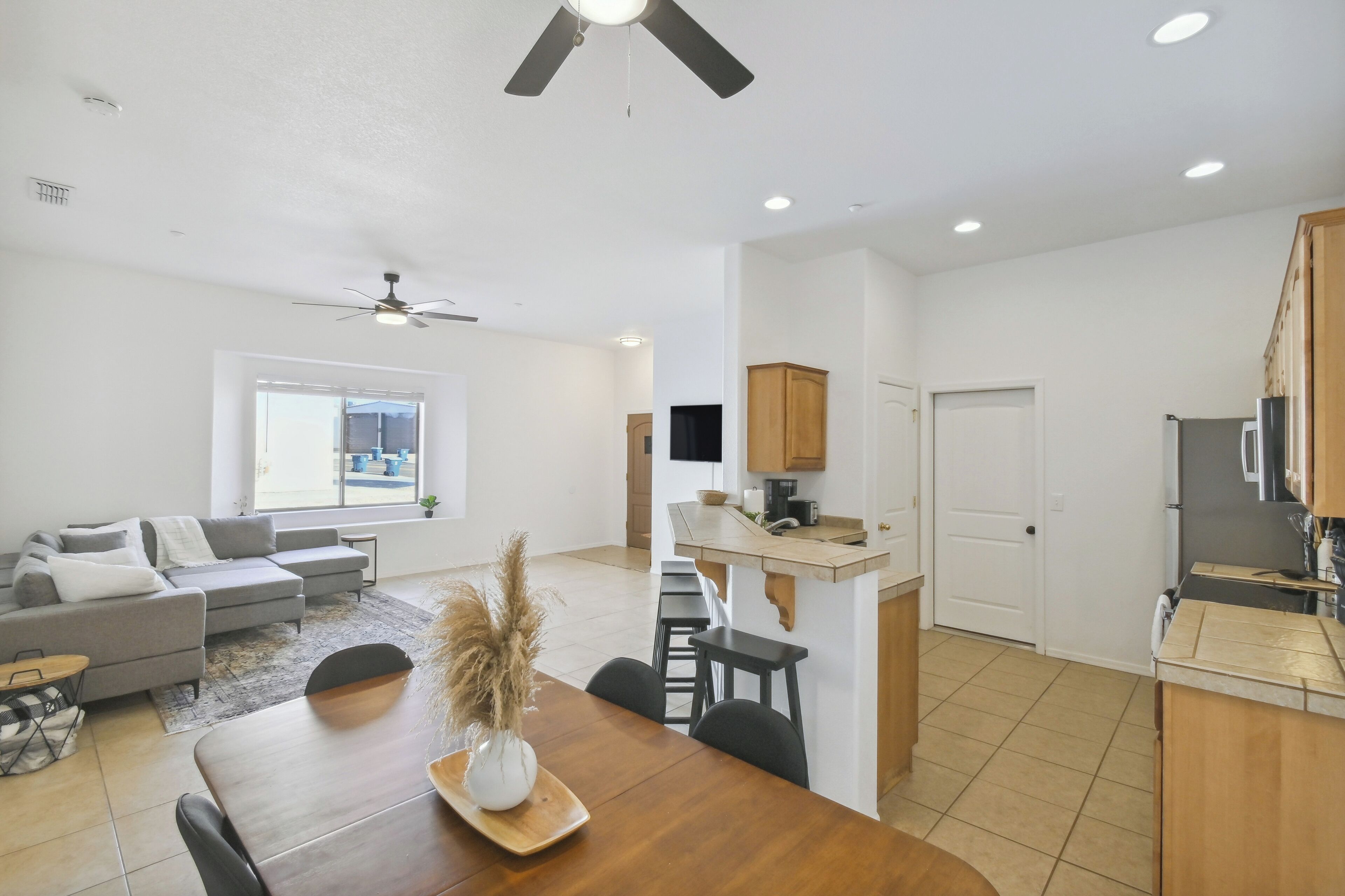 A modern living room with a grey sectional sofa, a wooden dining table, and a kitchen area with wooden cabinets and a built-in oven.