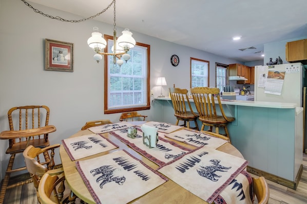 Comedor con mesa de madera, sillas y una lámpara de candelabro. Hay una zona de cocina con armarios de madera y un refrigerador.