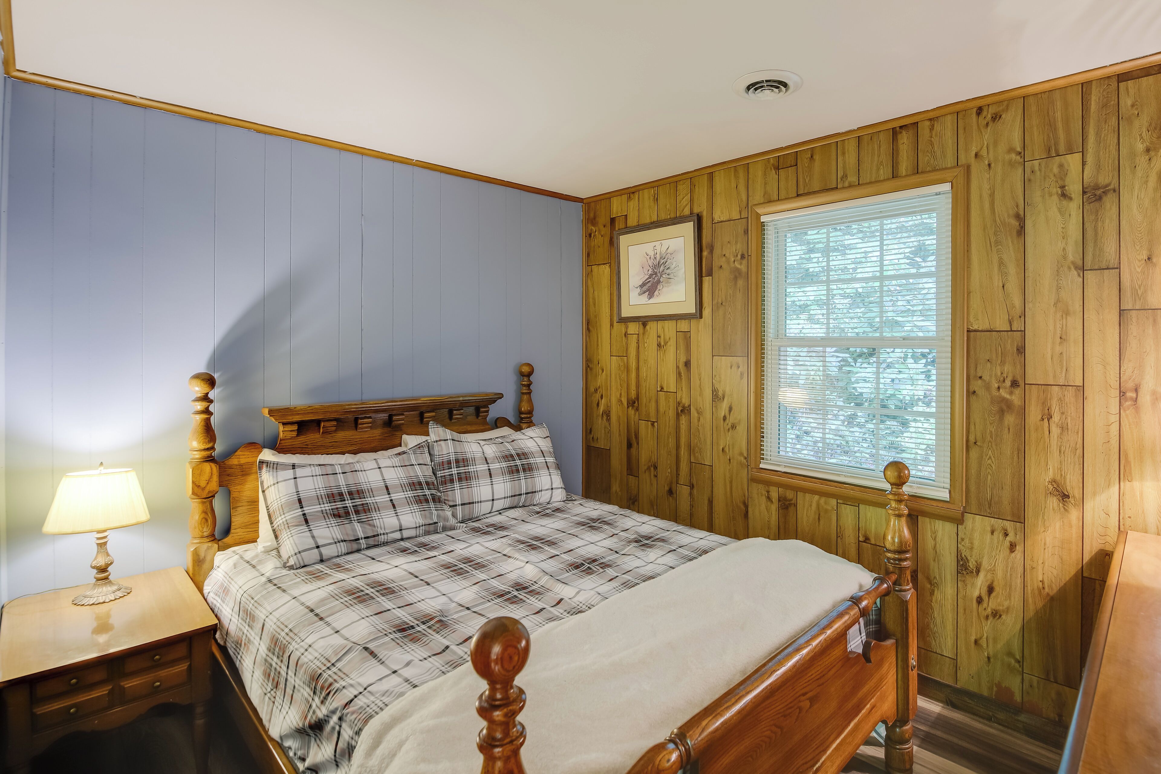Un dormitorio con cabecera de madera, colcha a cuadros, mesita de noche con lámpara y ventana con persianas.