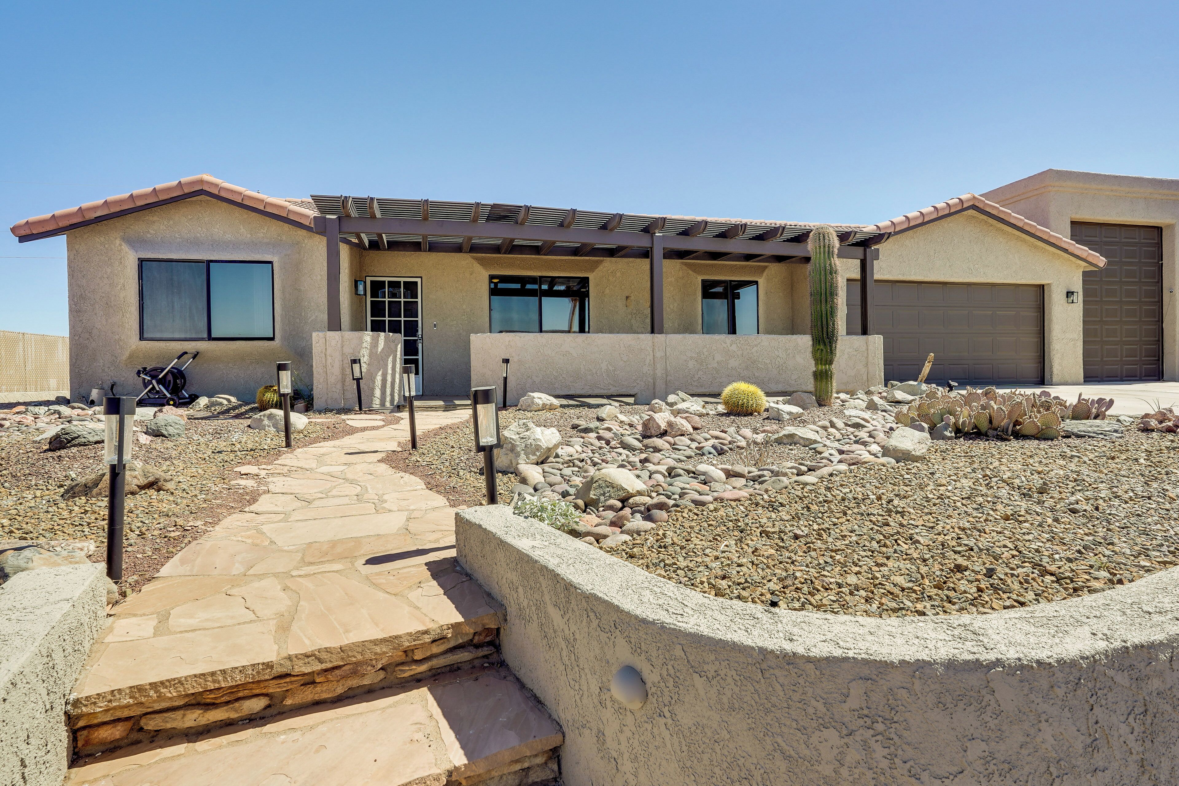 A modern house with a stone pathway leading to the entrance, a covered patio, and a desert garden with cacti and rocks.