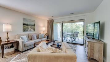 A modern living room with a sofa, coffee table, and TV. There is a balcony with a table and chairs.