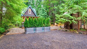 A cabin with a sloped roof, surrounded by dense forest and a gravel driveway. - Adorable A-frame Cabin, Steps to Lake Cushman! (Hoodsport)