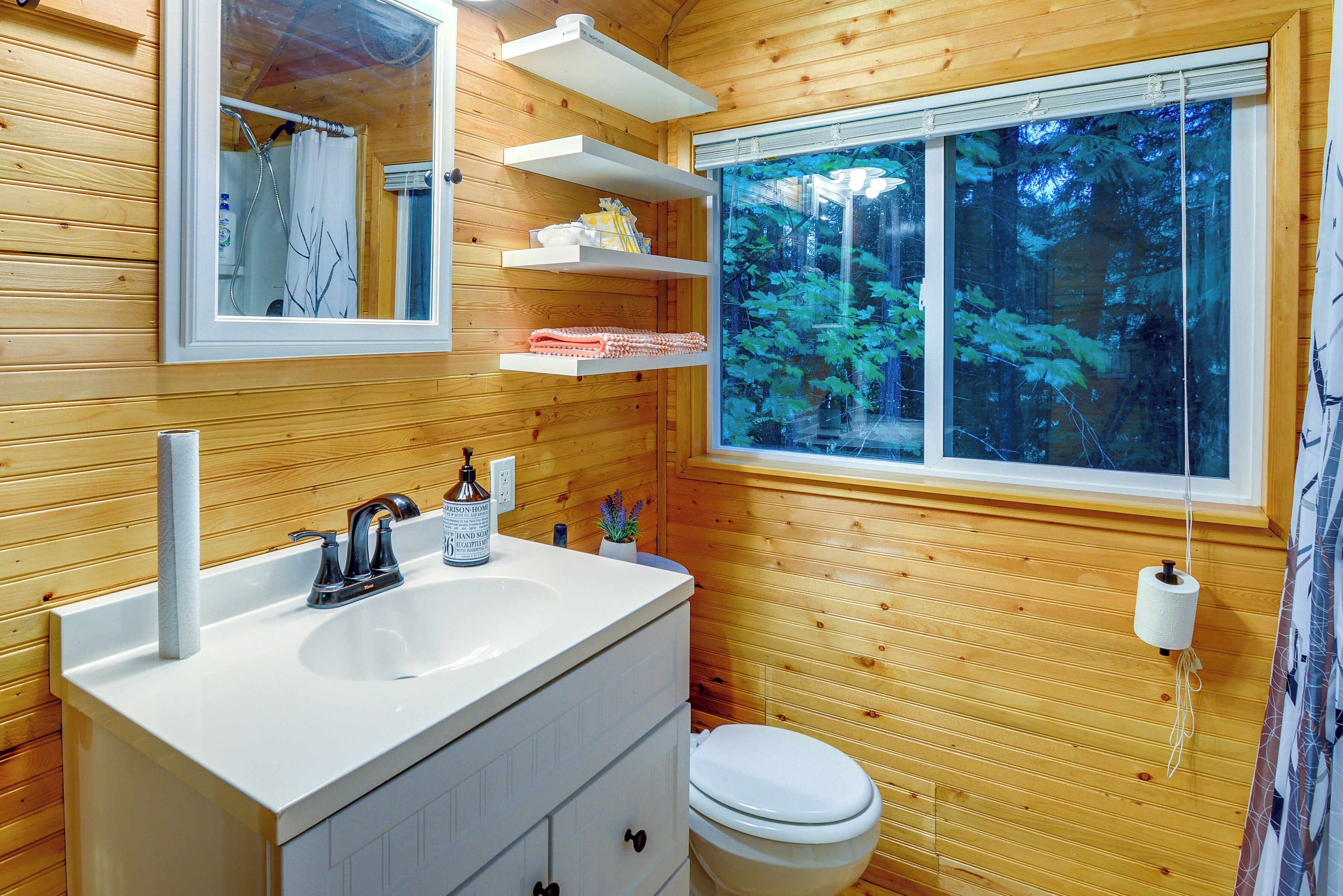 Une salle de bain rustique, avec des murs en bois, comprenant un lavabo blanc, un miroir et des toilettes. On y trouve des étagères au-dessus du lavabo et une fenêtre donnant sur les arbres.