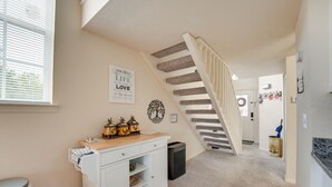 A hallway with a staircase, a white cabinet with a tray, a trash can, and a wall with a decorative wreath and a sign. - Branson Vacation Rental w/ Loft ~ 4 Mi to Downtown (Branson)