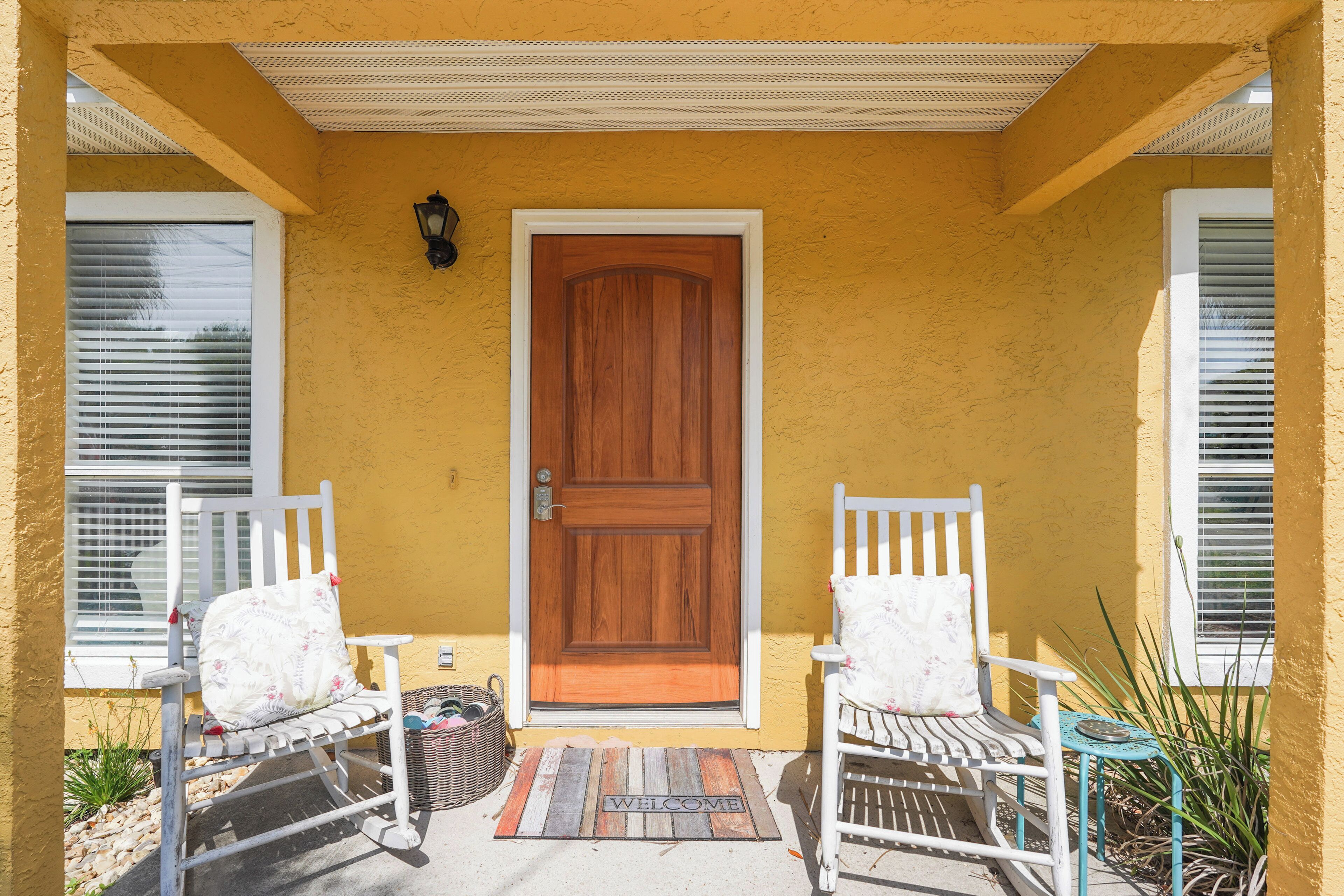 Una casa amarilla con puerta de madera, dos mecedoras blancas y un felpudo.