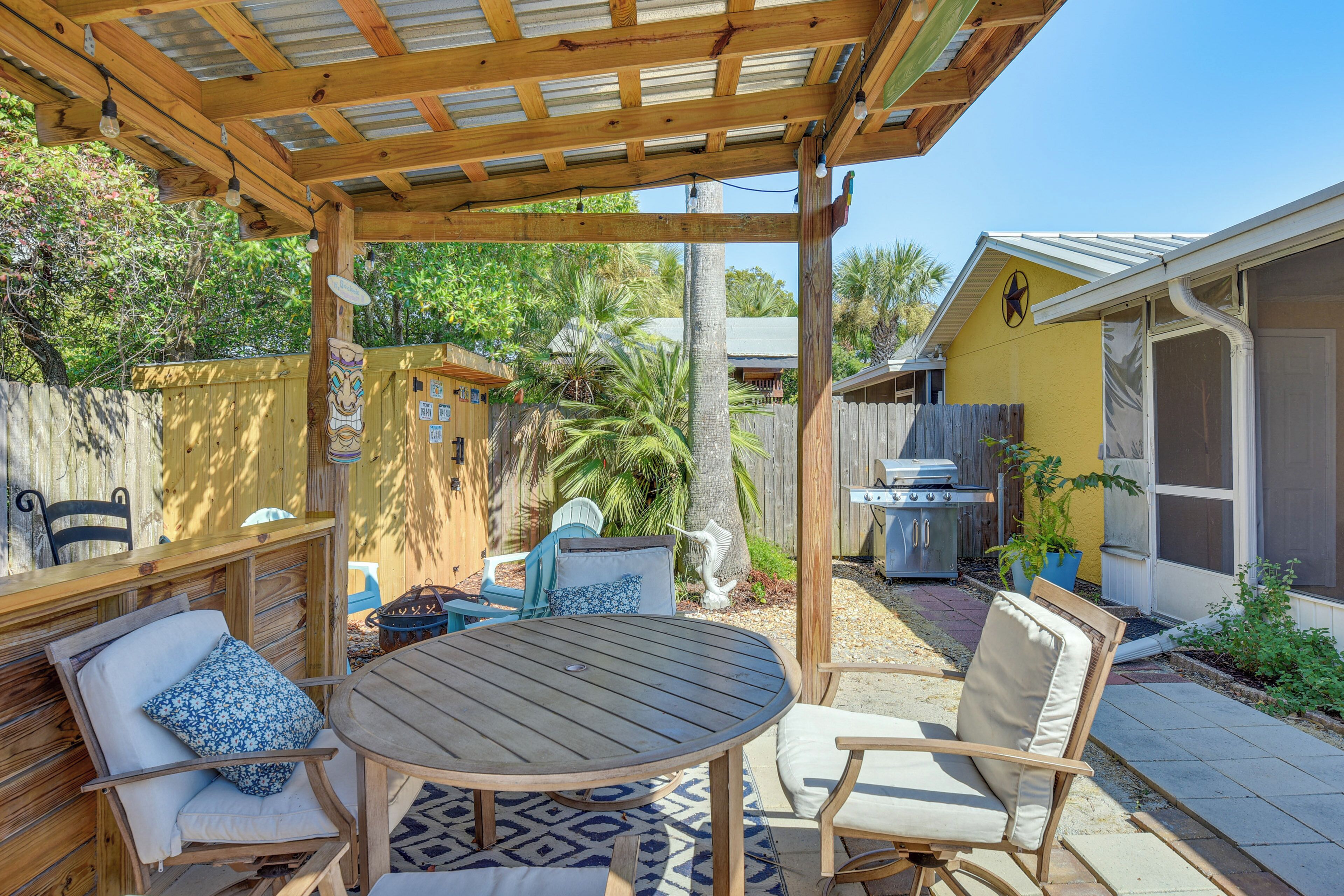 Un patio techado con mesa y sillas de madera, una parrilla y un jardín trasero cercado.