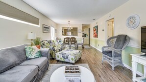 A spacious living room with a grey sofa, a patterned ottoman, and a wicker chair. A chandelier hangs from the ceiling, and a clock is mounted on the wall. - Breezy PCB Vacation Rental w/ Lanai, Walk to Ocean (Panama City Beach)