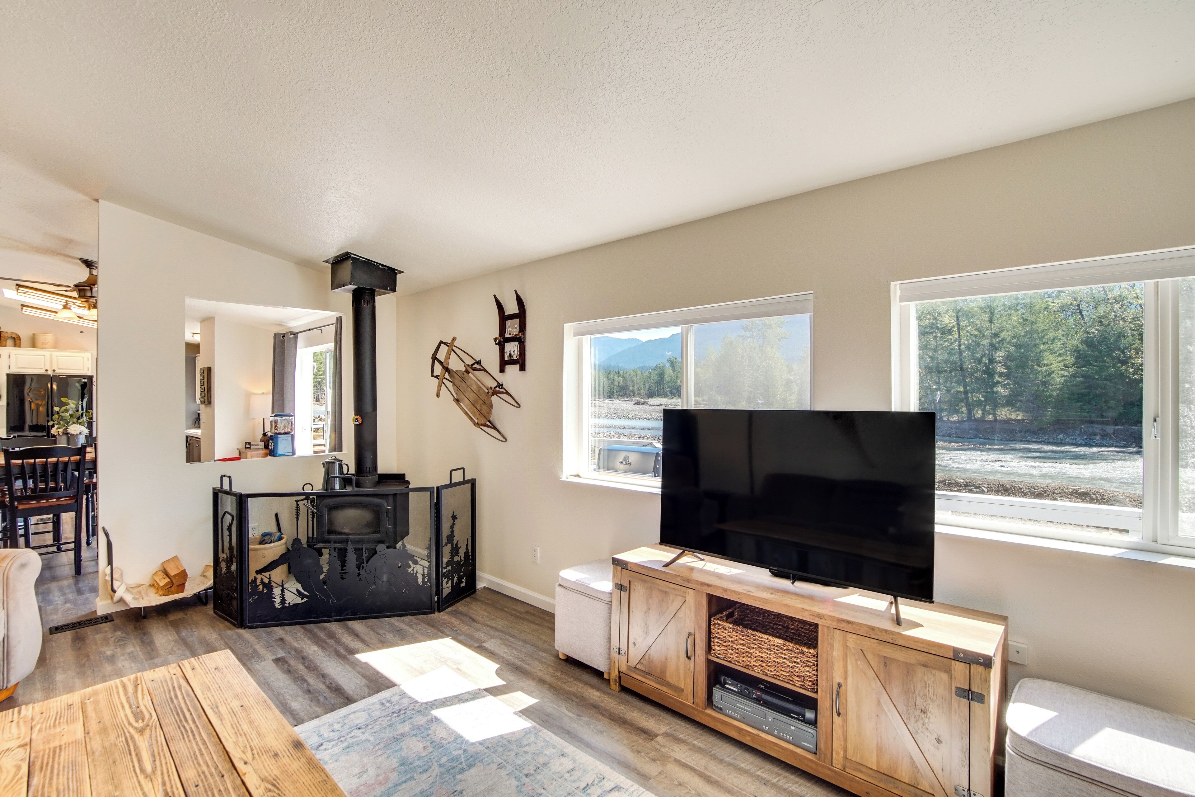 A modern living room with a fireplace, a large flat-screen TV, and a view of a forest through the windows.