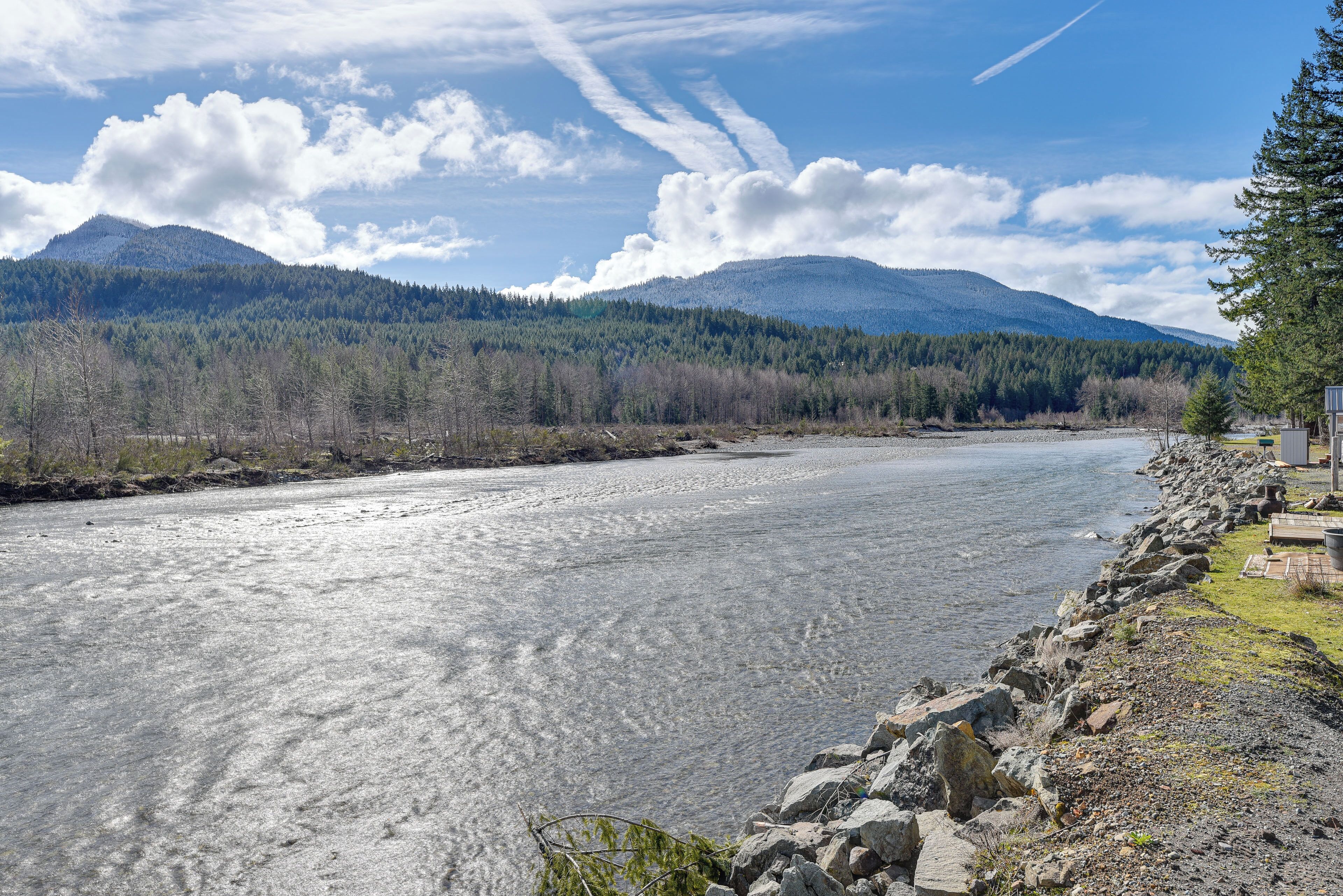 A river with a rocky shoreline, a forested mountain in the background, and a clear sky.