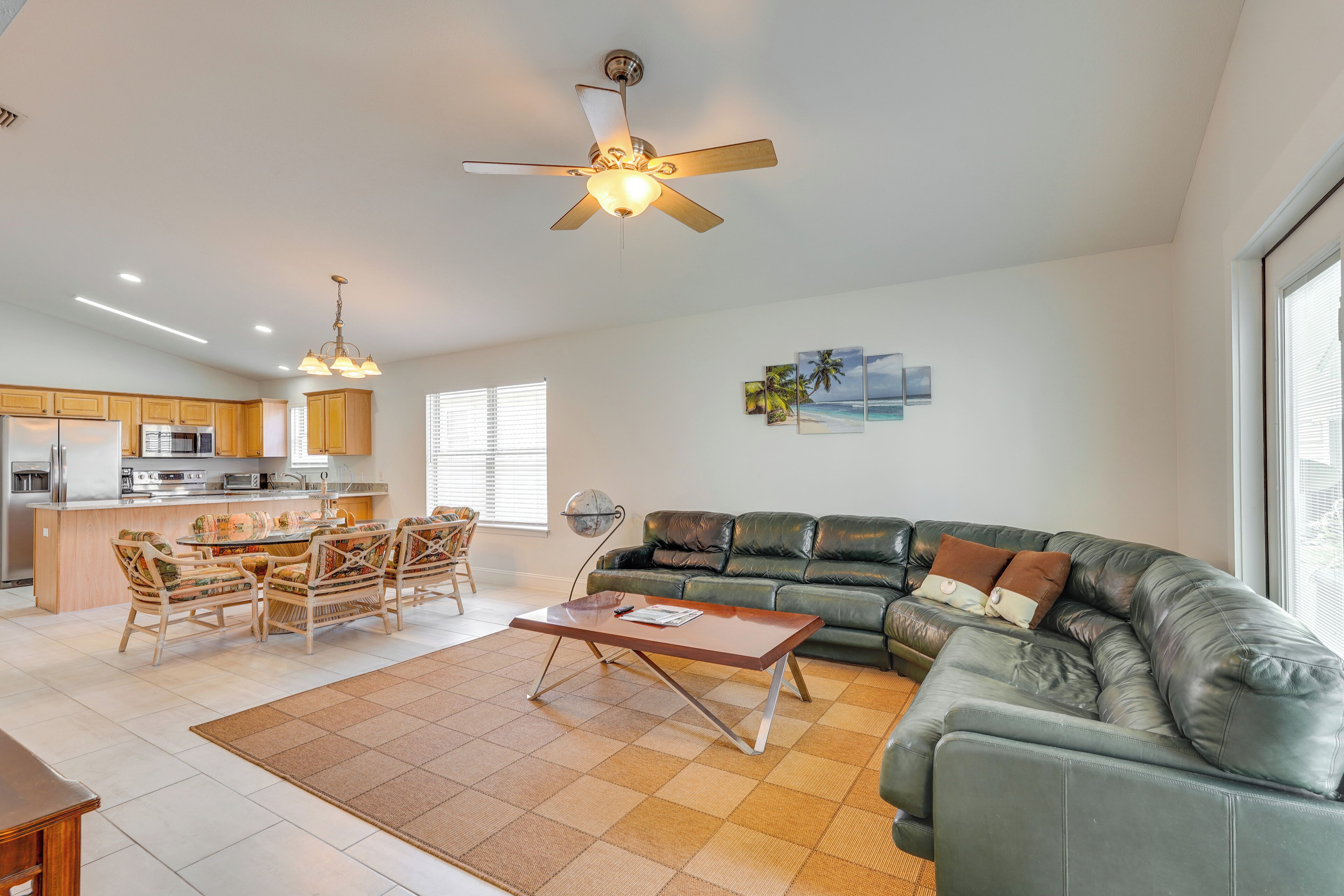 A spacious living area with a sofa, coffee table, and dining set. There is a ceiling fan, a kitchen area with cabinets, and a large window providing natural light.