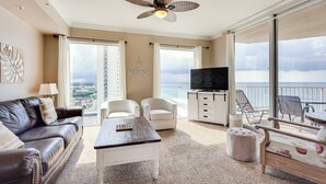A spacious living room with a leather sofa, a coffee table, and a TV. There are two white armchairs, a small ottoman, and a balcony with a view of the ocean. - Panama City Beach Condo: Balcony w/ Ocean Views! (Panama City Beach)