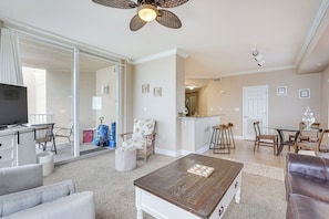 A spacious living area with a large flat-screen TV, a wooden coffee table, and a leather sofa. There is a sliding glass door leading to a balcony, and a kitchen area is visible in the background. - Panama City Beach Condo: Balcony w/ Ocean Views! (Panama City Beach)