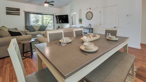 Un coin repas avec une longue table dressée pour quatre personnes, un ventilateur de plafond et un téléviseur en arrière-plan.