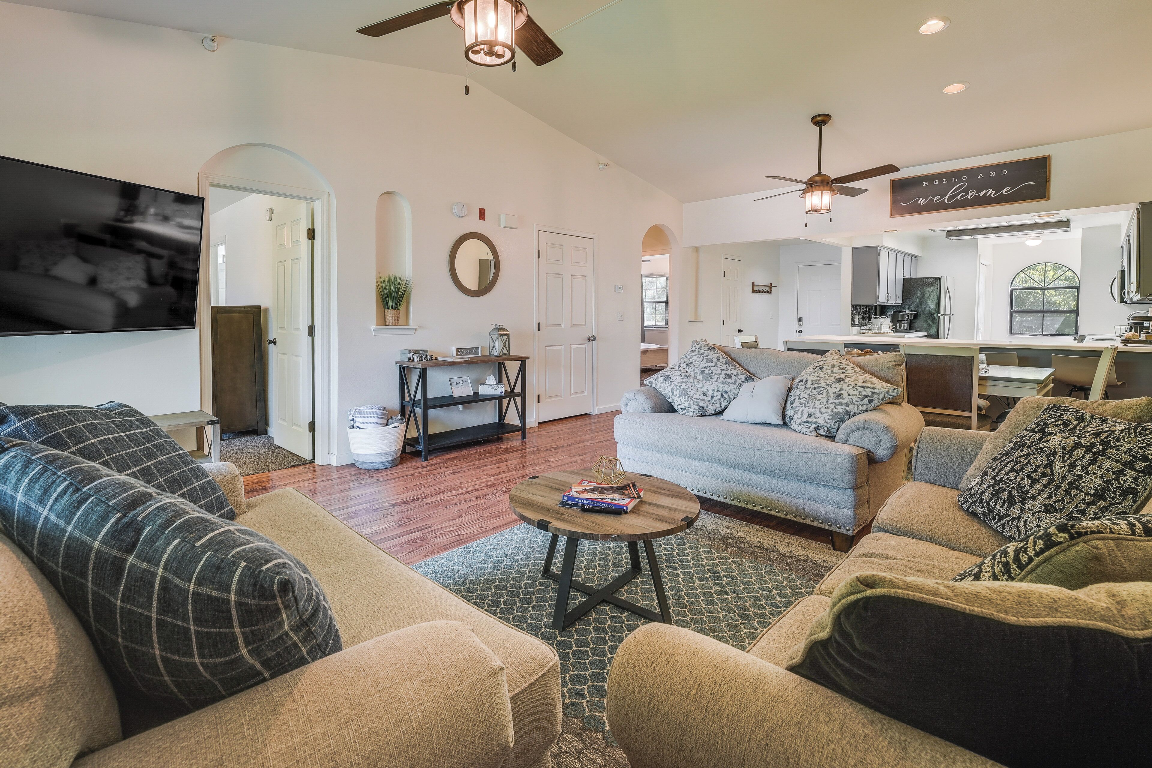 A spacious living room with a large flat-screen TV, a ceiling fan, and a kitchen area visible through an open doorway.