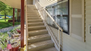 Un escalier menant à un porche couvert doté d’une balustrade, une fenêtre avec des stores et un petit jardin agrémenté de plantes et d’une mangeoire à oiseaux.