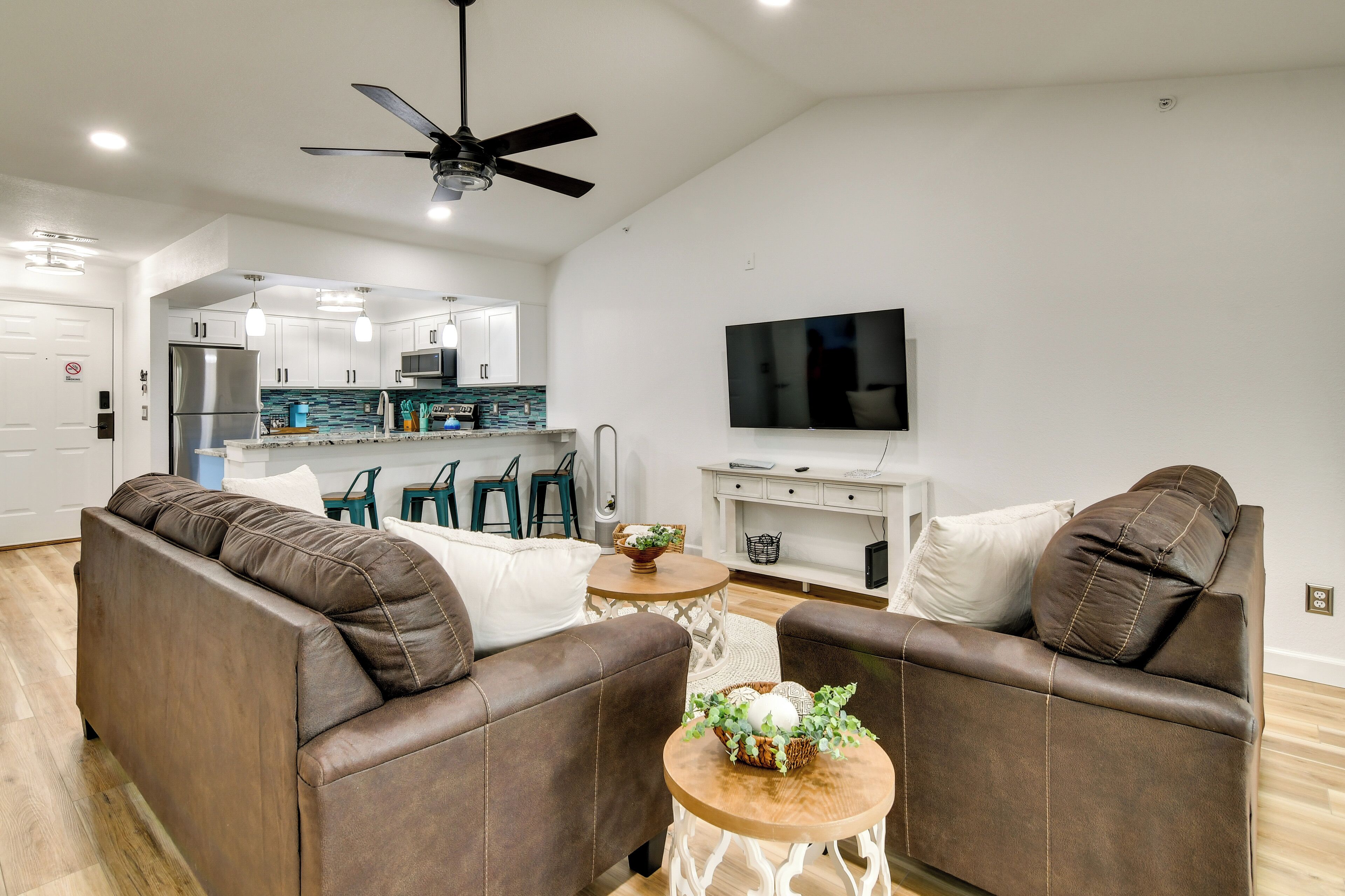 A modern living room with a brown leather sofa, a flat-screen TV mounted on the wall, and a ceiling fan.