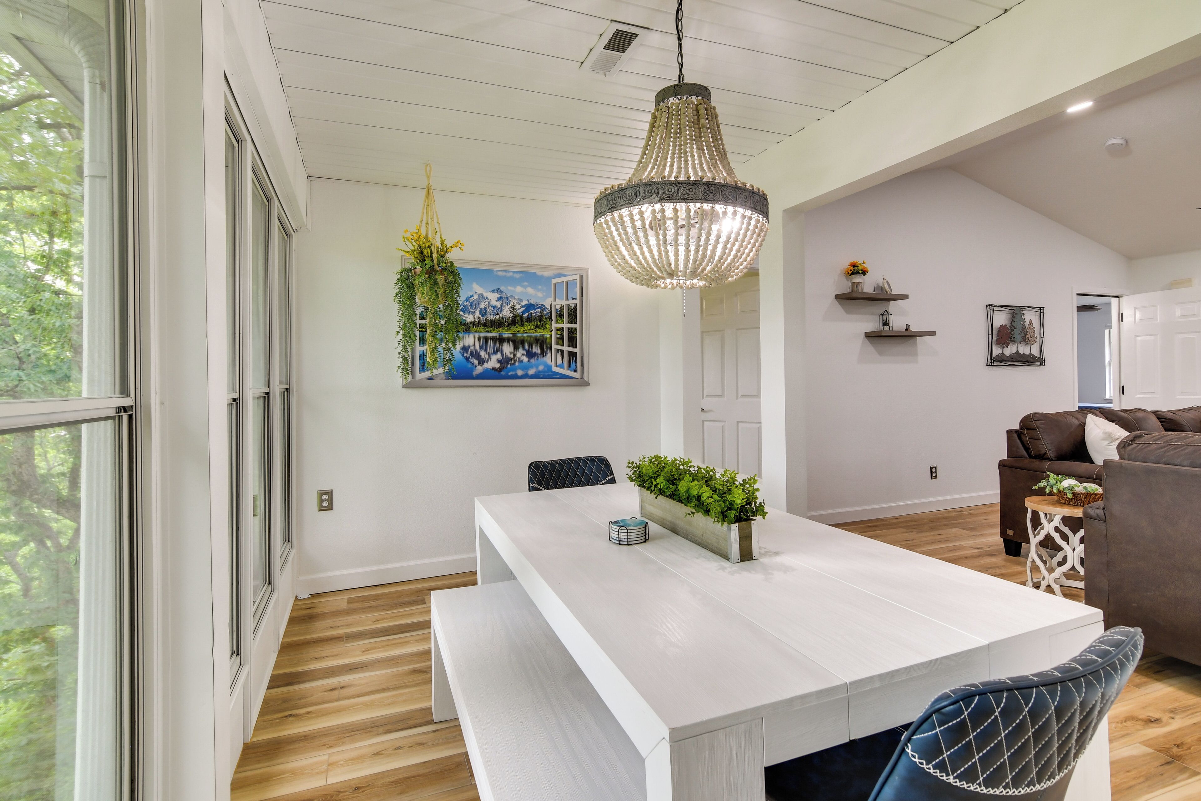 A modern dining area with a white table, bench seating, a chandelier, and a painting of a mountain landscape.