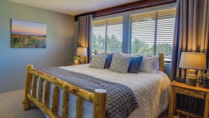 Une chambre avec un lit en bois, une grande fenêtre avec des stores, un tableau au mur et deux tables de chevet avec des lampes.