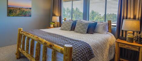 A bedroom with a wooden bed frame, a large window with blinds, a painting on the wall, and two bedside tables with lamps.