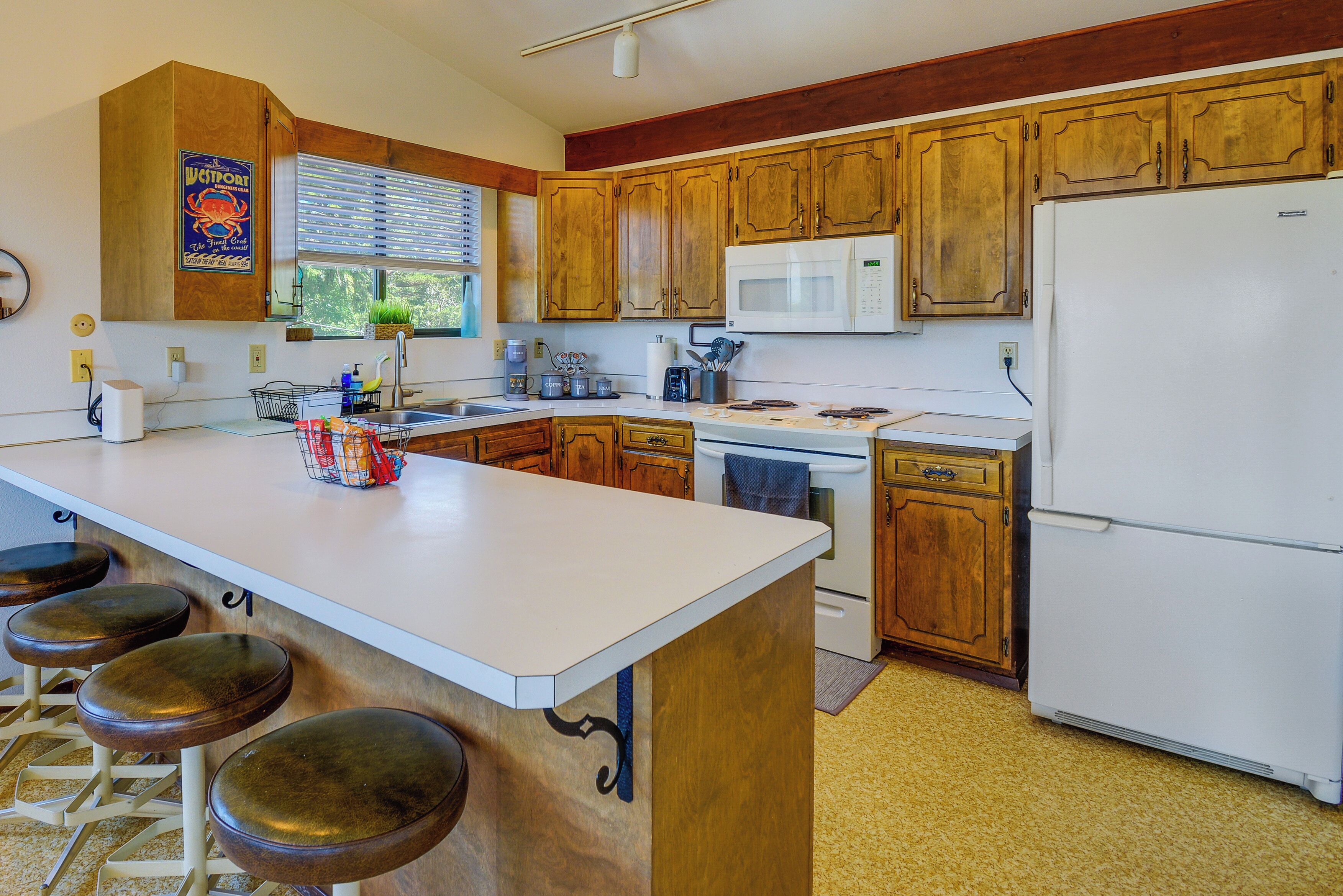 A kitchen with wooden cabinets, a white countertop, a sink, a microwave, a refrigerator, and bar stools.