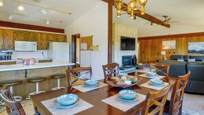 On aperçoit un coin repas avec une table en bois dressée pour six personnes, entourée de chaises en bois. Au fond, on distingue une cuisine avec des placards en bois et un four à micro-ondes.