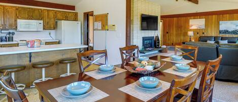 Uma sala de jantar com mesa de madeira posta para seis pessoas, rodeada por cadeiras de madeira. Uma cozinha com armários de madeira e um micro-ondas é visível ao fundo.