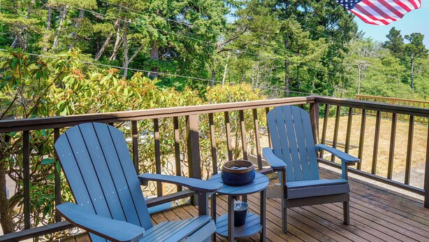 Une terrasse en bois avec deux fauteuils Adirondack et une petite table, entourée d’arbres et offrant une vue sur la forêt.