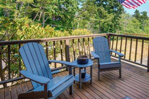 Une terrasse en bois avec deux fauteuils Adirondack et une petite table, entourée d’arbres et offrant une vue sur la forêt.